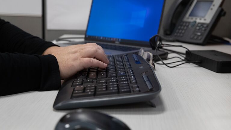 Worker-typing-to-a-keyboard-with-laptop-and-phone-in-the-background